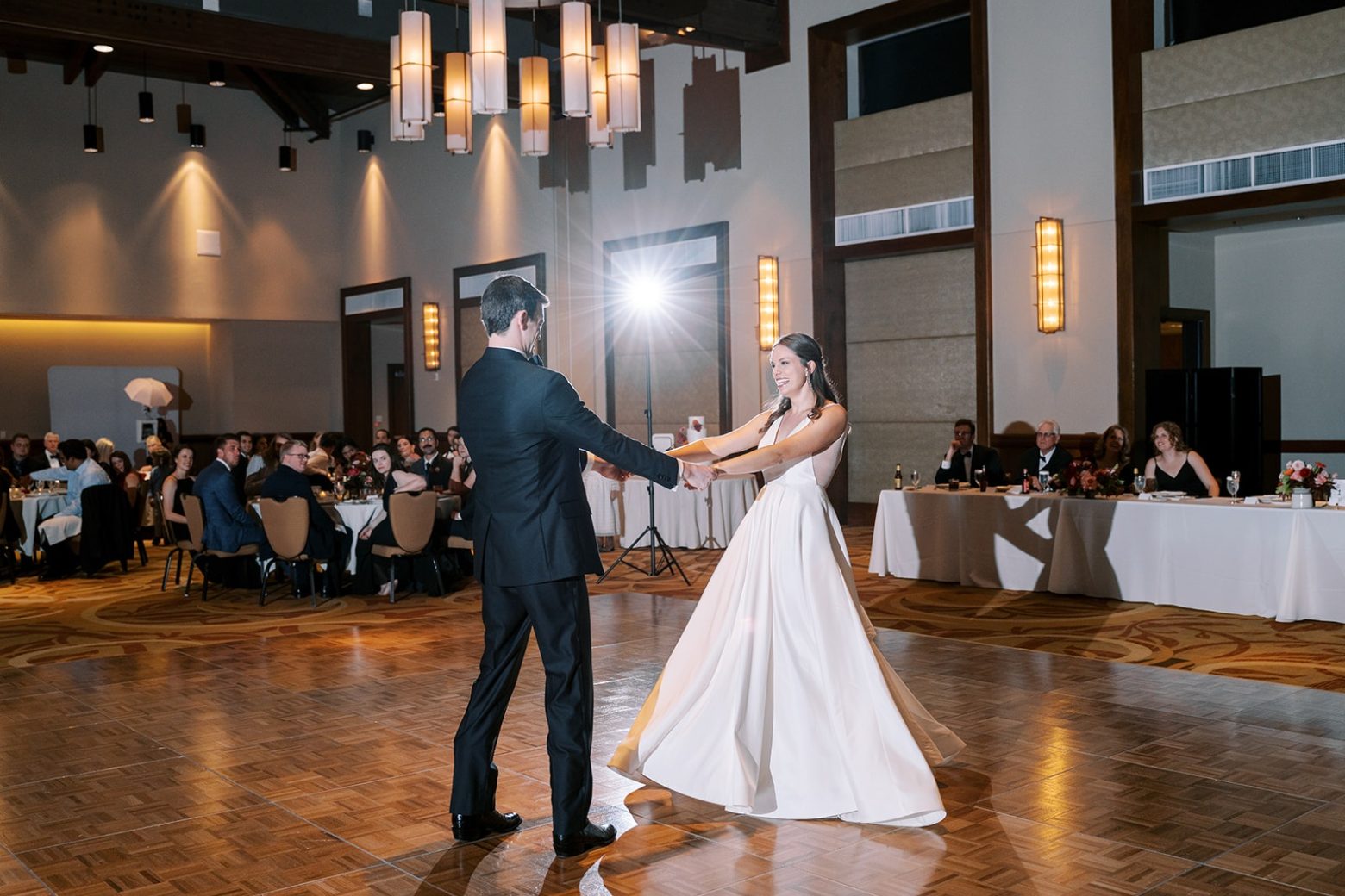 A large room with a high ceiling, parquet floor, and chandelier, where a couple is dancing.