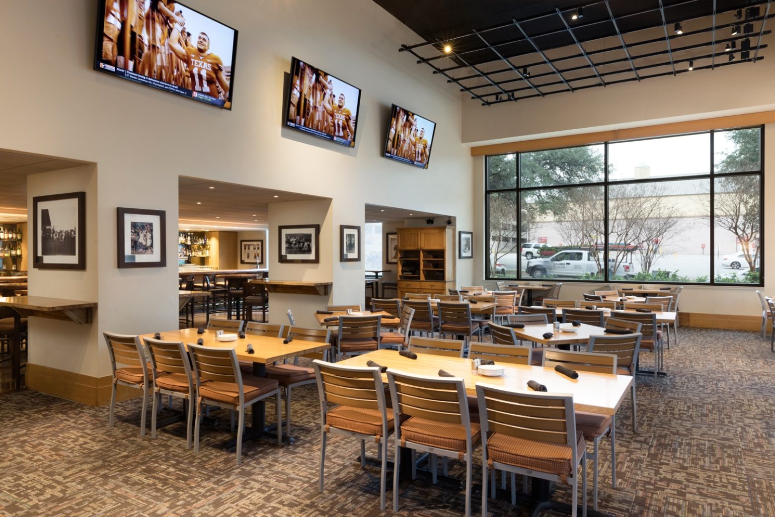 A dining area with tables and chairs, TV screens on the wall, and a large window.