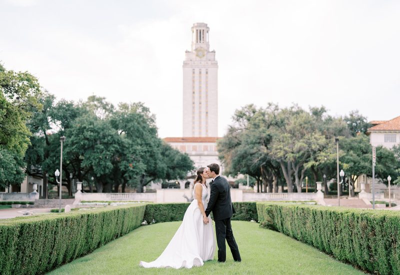 A couple in wedding attire stands in a manicured garden with a tall clock tower in the background.
