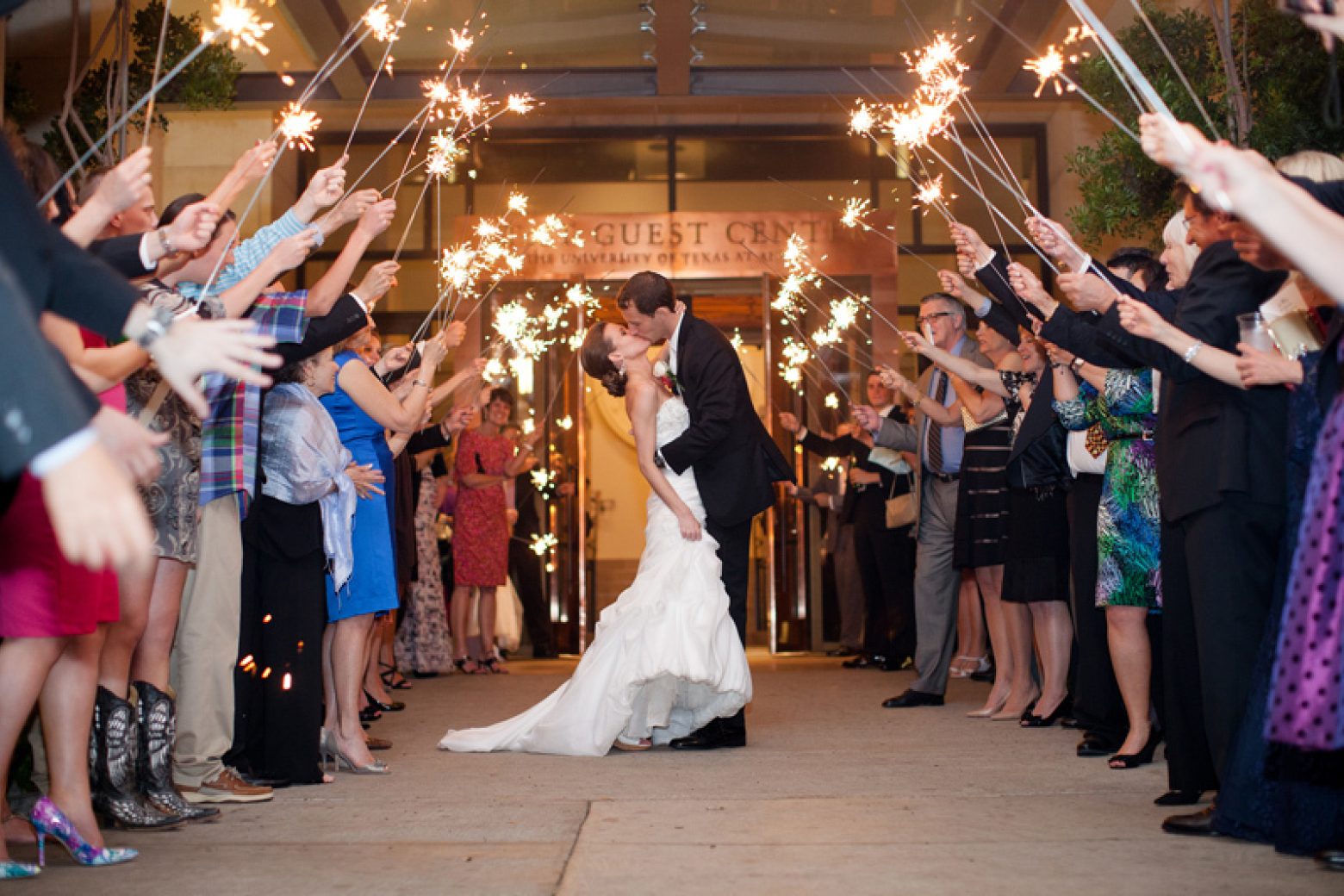 A wedding couple is surrounded by people holding sparklers in front of a building entrance.