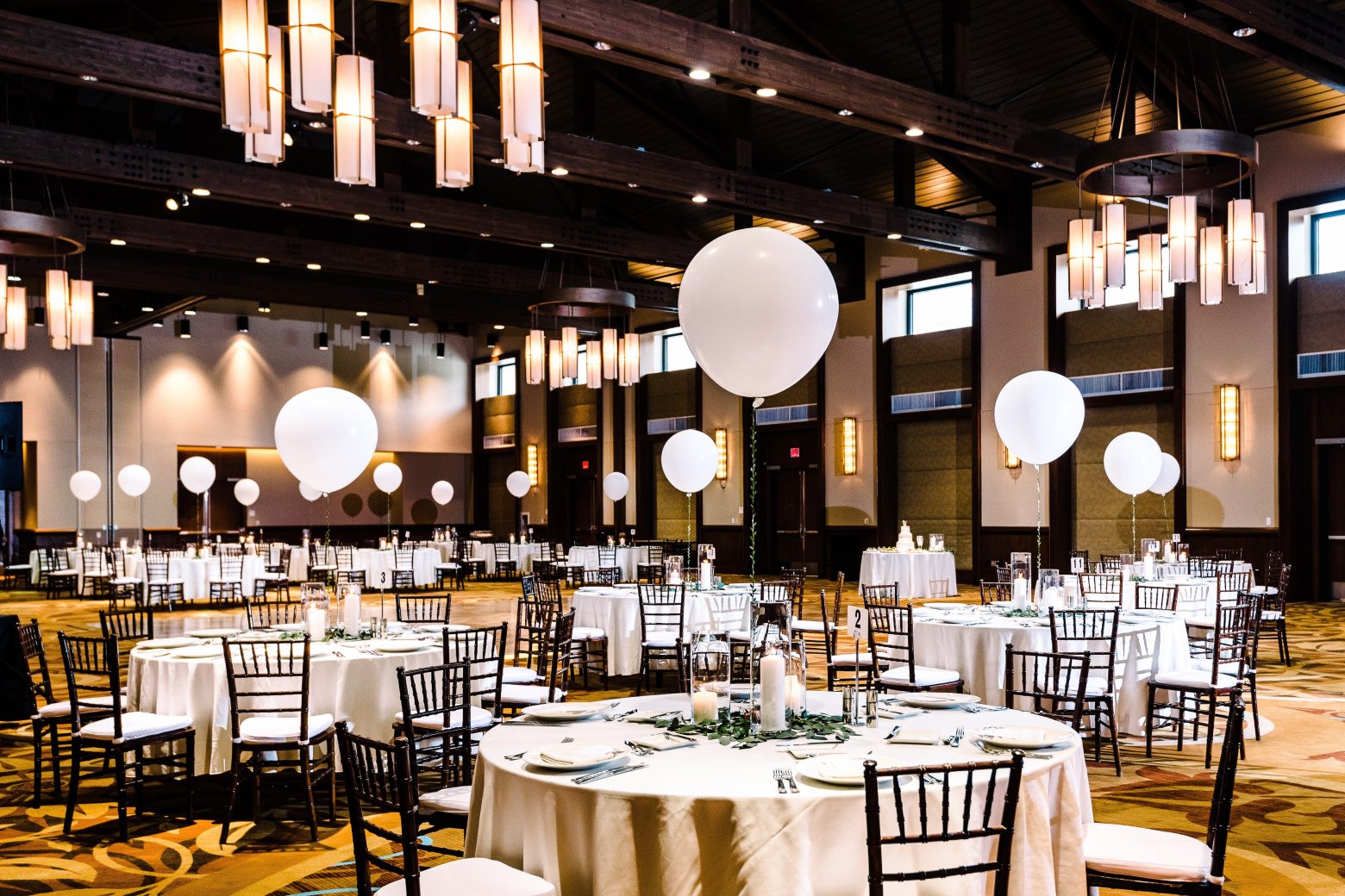 A large, elegantly decorated meeting room with round tables, white tablecloths, black chairs, and white balloons.