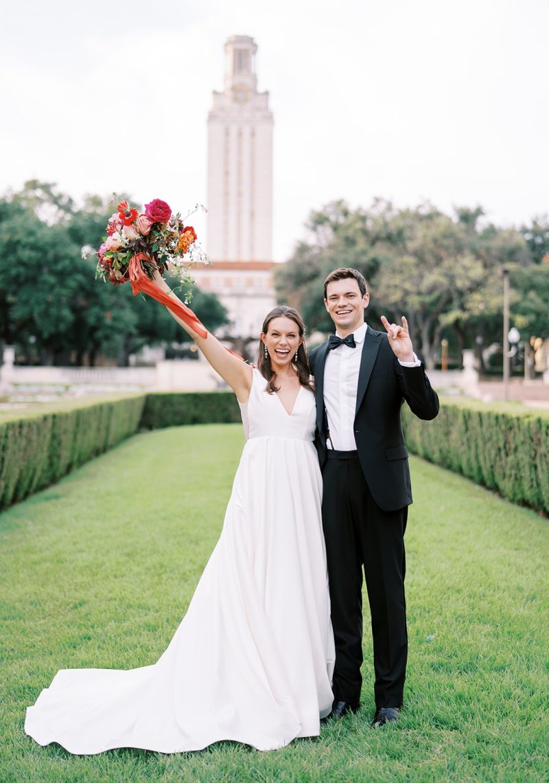 A bride and groom stand in a grassy area with a tower in the background.