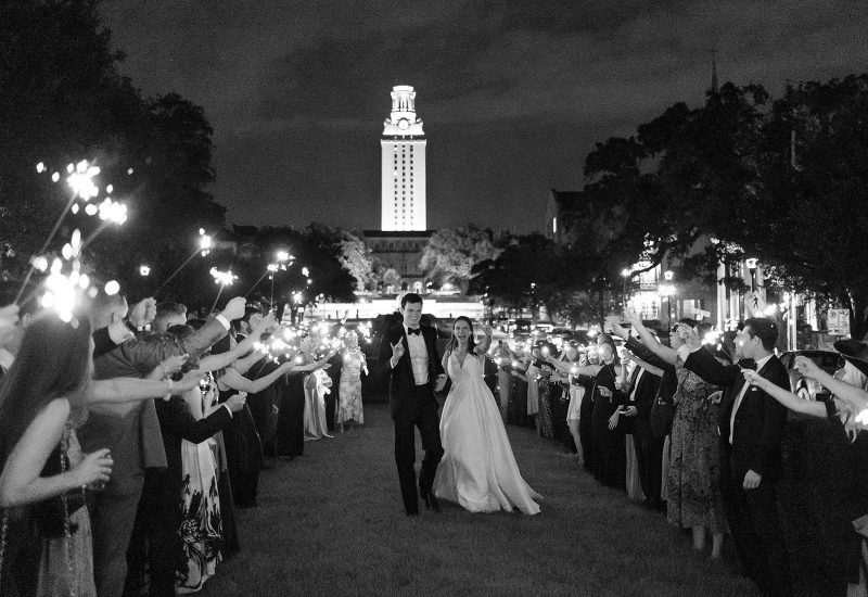 a black and white photo of a couple walking down a pathway surrounded by people holding sparklers with a tower in the background