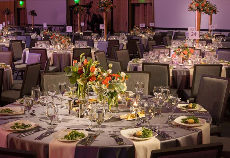 A large, elegantly set dining room with round tables, gray chairs, and flower arrangements.