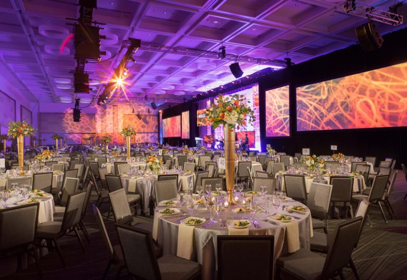 A large meeting room with round tables, white tablecloths, gray chairs, and flower centerpieces, with large screens on the walls and purple lighting.