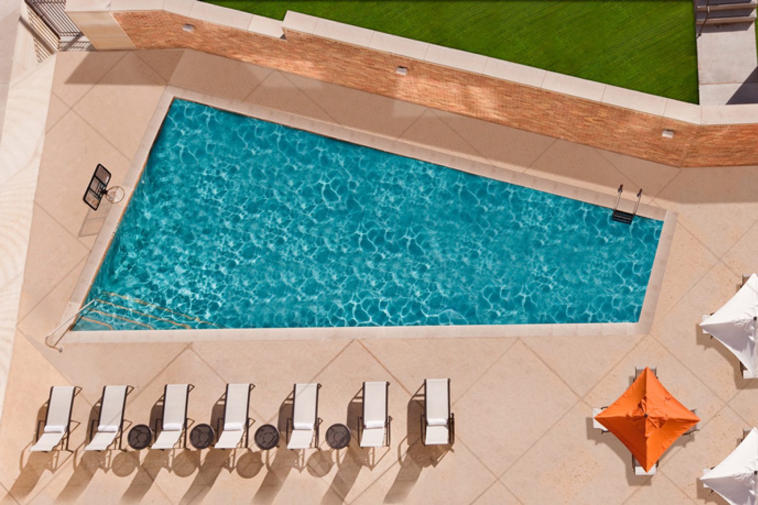 An aerial view of a rectangular pool with a row of white lounge chairs and an orange umbrella.